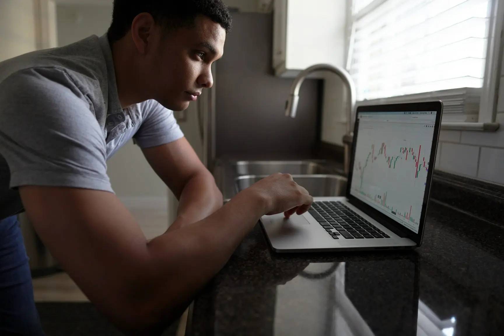 A man leans over a kitchen counter, intently analyzing fluctuating stock market charts on his laptop in natural daylight.